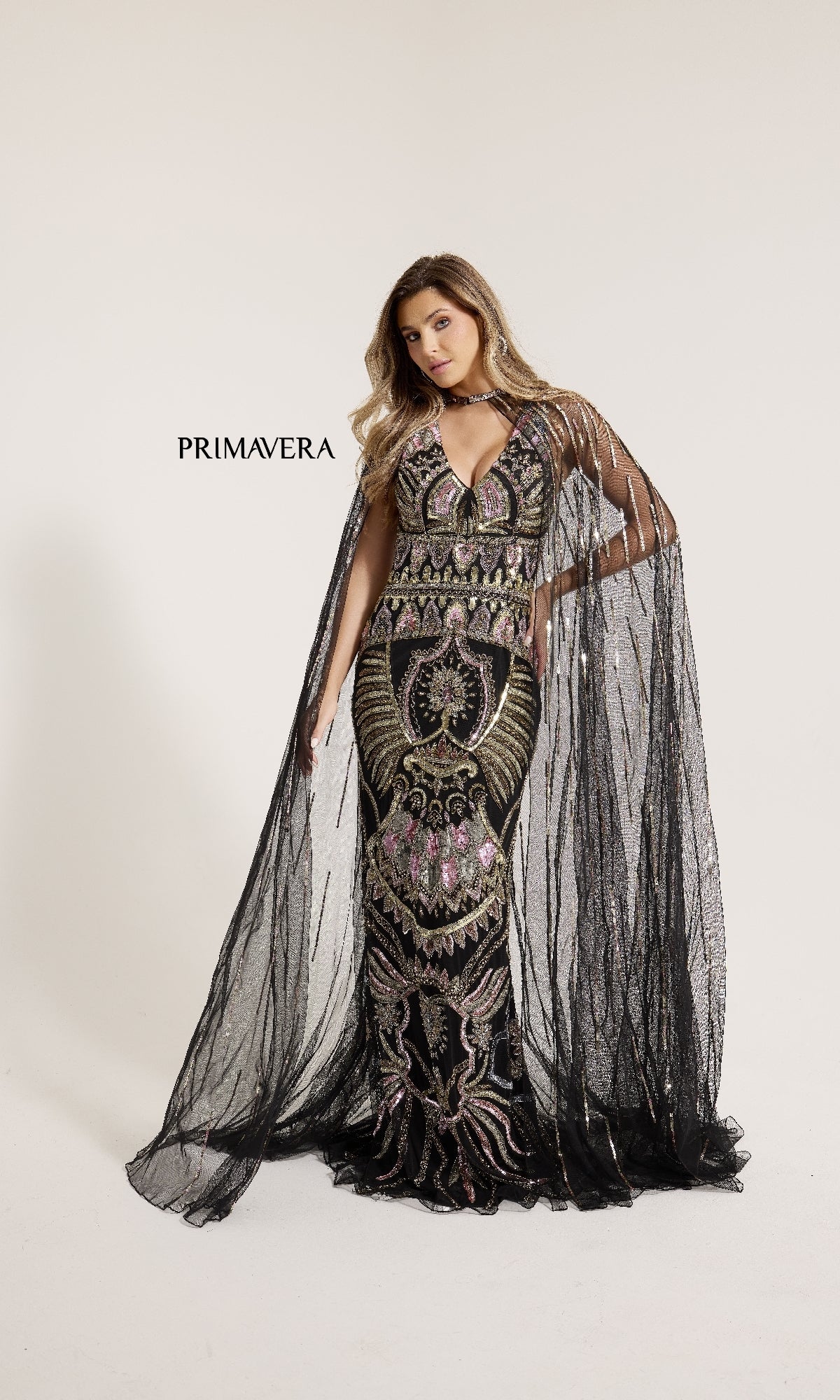 A woman models the Primavera 12177, a long formal black and gold gown with an ornate design and sheer floor-length cape, standing against a white background. The word